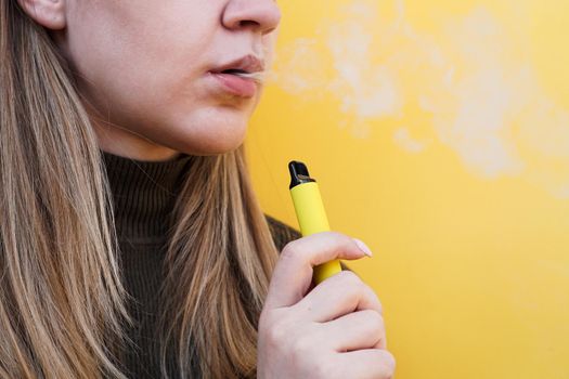 Close up of a young girl smokes a disposable electronic cigarette and exhales smoke. Bright yellow background. Alternative to regular cigarettes and vape