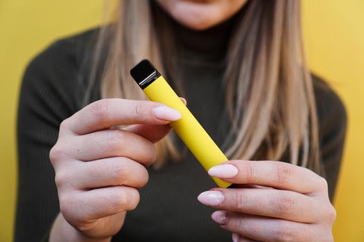 Yellow disposable electronic cigarette in a female hand. Bright yellow background. Melon, pineapple or lemon flavored vape