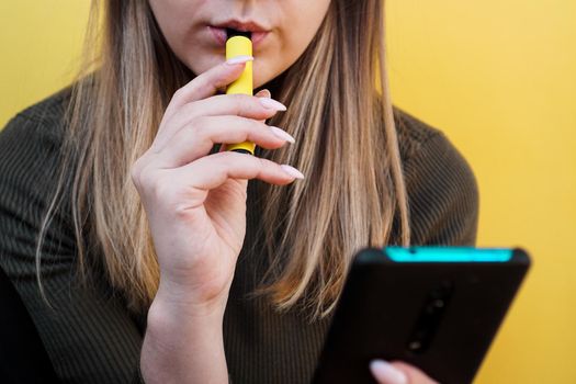 Close up of a young girl smokes a disposable electronic cigarette. Bright yellow background. Alternative to regular cigarettes and vape