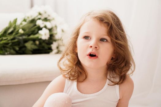 Little three-year-old girl with a surprised face. Cute baby at home. Light interior and white flowers on the background