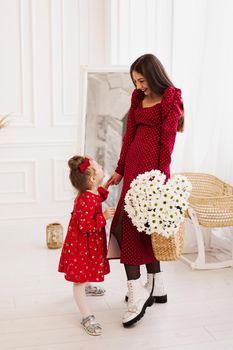 Mom and little daughter in red dresses in a bright bedroom in a Scandinavian style. Happy family. Mom holds a bouquet of daisies - spring photo