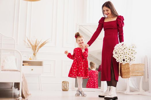 Mom and little daughter in red dresses in a bright bedroom in a Scandinavian style. Happy family. Mom holds a bouquet of daisies - spring photo