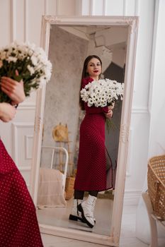 Beautiful brunette in a red dress with a bouquet of daisies looks in the mirror at home. Spring photo. Full-length photo