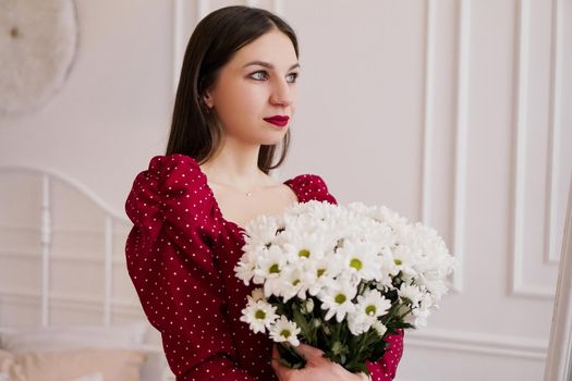 Beautiful brunette in a red dress with a bouquet of daisies at home. Spring photo.