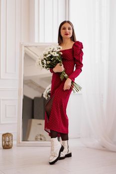 Beautiful brunette in a red dress with a bouquet of daisies at home. Spring photo. Full-length photo