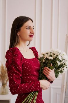 Beautiful brunette in a red dress with a bouquet of daisies at home. Spring photo.