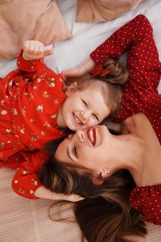 Mom and her little daughter lie on the bed facing each other. They are happy. Both are wearing red dresses.