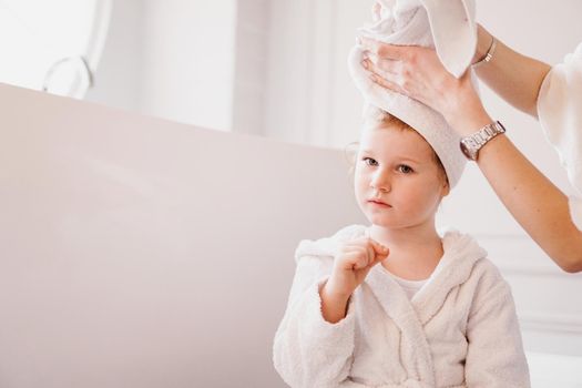 Mom puts a towel on her daughter's hair. Little daughter in a white bathrobe in a bright bathroom.