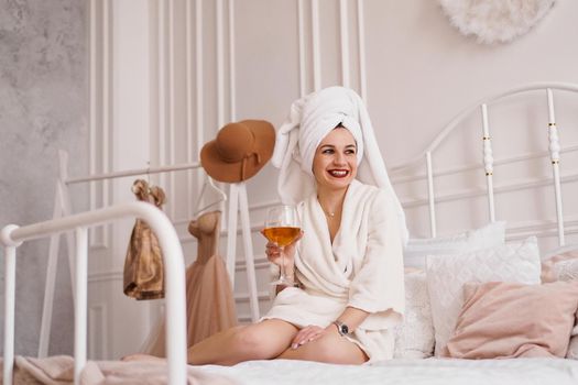 Beautiful woman in a white robe and with a towel on her hair. She is holding a glass of white wine. Light Scandinavian bedroom interior.