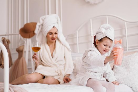 Mother and daughter in the bedroom in bathrobes. The mother drinks a glass of white wine, and the daughter drinks juice from a glass. A wonderful holiday with the family