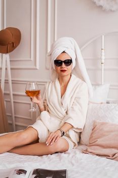 Beautiful woman in a white bathrobe, towel and sunglasses. She is holding a glass of wine. The concept of relaxation or morning of the bride. Scandinavian style hotel room