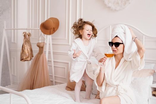 Mother and daughter in the bedroom in bathrobes. Happy daughter is jumping on the bed. Mom is drinking a glass of white wine. Funny motherhood fatigue concept