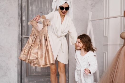 Happy mother and daughter are choosing a dress in the wardrobe. The mother is wearing a white bathrobe. They are going to the party.