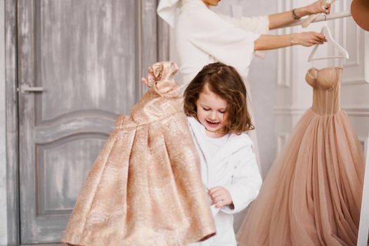 The little girl chose a beige dress in the wardrobe and is going to wear it. Happy child. Mom in the background