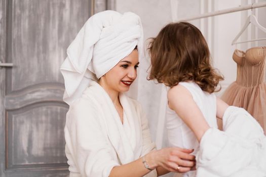 Attractive mother in a white bathrobe is dressing her little daughter. Against the background of a blurred hanger with a beige dress. Wardrobe concept