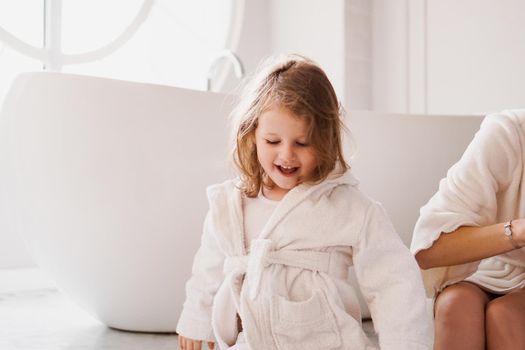 Smiling little girl in a white bathrobe after a bath. White cozy interior. Hygiene and baby fashion concept