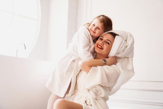 Little daughter hugs mom. Mother and daughter in white coats in a light bathroom. Hygiene, beauty and happy family concept