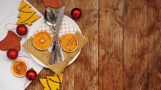 Overhead view of Christmas table setting over wooden table with copy space. White plate with dry oranges, red balls and wooden christmas figurines