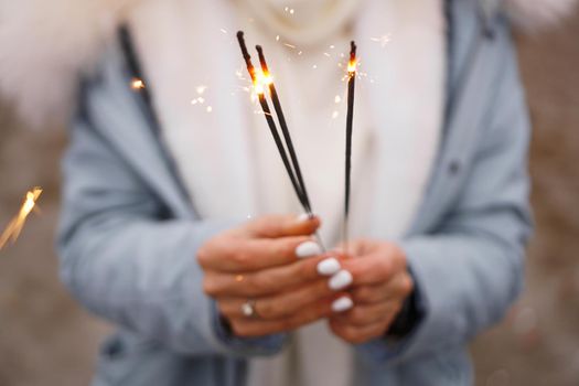 Female hands are holding burning sparklers. Woman outdoors in winter. Blurred background