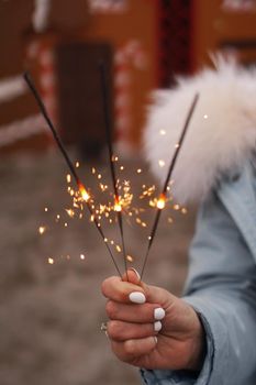 Female hands are holding burning sparklers. Woman outdoors in winter. Blurred background