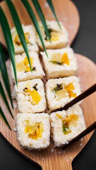 Dessert sushi. Sweet kiwi, pineapple sushi rolls. Sushi on a wooden tray on black background with tropical leaf. Vertical photo. Holding a sweet roll