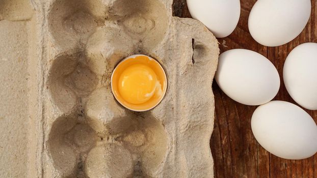 White eggs carton and cracked egg half with yolk top view on wooden background. Easter and healthy food breakfast cooking concept