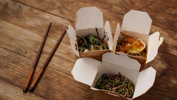 Noodles with pork and vegetables in take-out box on wooden table. Asian food delivery. Food in paper containers on wooden background