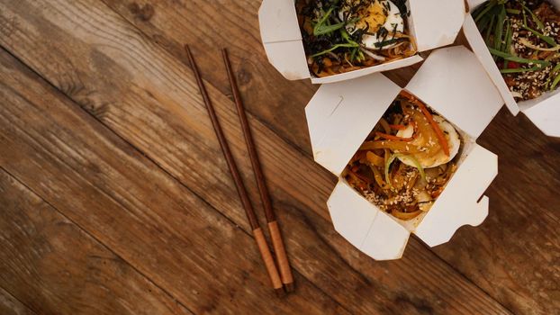 Noodles with pork and vegetables in take-out box on wooden table. Asian food delivery. Food in paper containers on wooden background