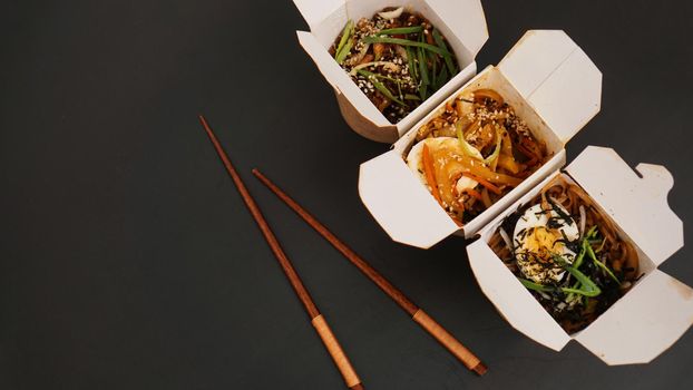 Noodles with pork and vegetables in take-out box on black table. Asian food delivery. Food in paper containers on black background