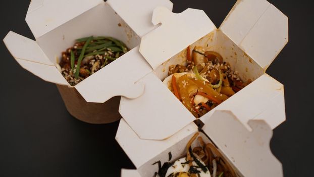 Noodles with pork and vegetables in take-out box on black table. Asian food delivery. Food in paper containers on black background