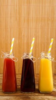 Three multicolored drinks in the bottles on a wooden bamboo background. Bottled paper straws. Vertical photo. Full length bottles