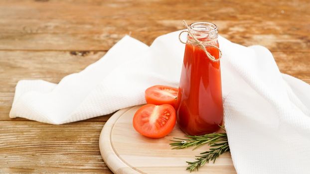 Tomato juice in glass bottle, fresh tomatoes and sprigs of rosemary on wooden cutting board and white towel