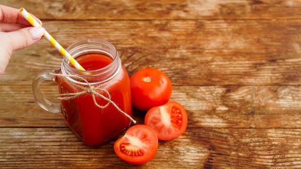 Female hand adding paper straw to glass of tomato juice on wooden table. Fresh tomato juice and chopped tomatoes on wooden board. Vertical photo