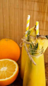 Two bottles of tropical juice with paper straws. Oranges, pineapple and rosemary for decoration. Wooden background. Vertical photo