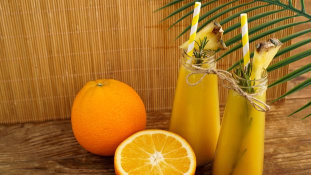 Two bottles of tropical juice with paper straws. Oranges, pineapple and rosemary for decoration. Wooden background.