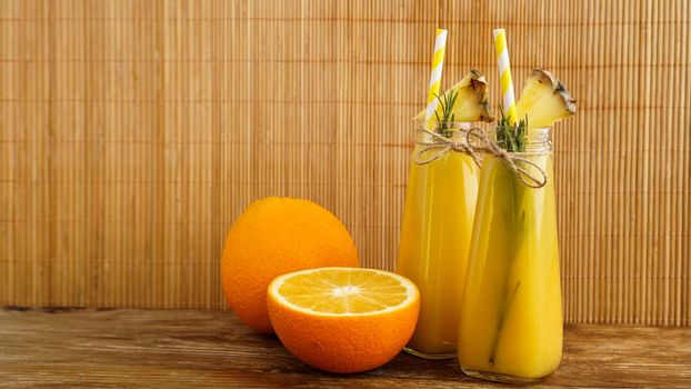 Two bottles of tropical juice with paper straws. Oranges, pineapple and rosemary for decoration. Wooden background.