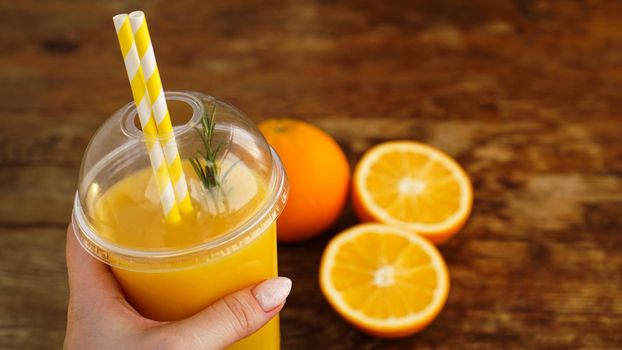 Girl hand is holding plastic glass with orange juice, wooden background with orange slices