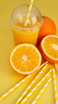 Orange juice in fast food closed cup with tube on yellow background. Sliced orange and yellow paper straws for a drink. Vertical photo