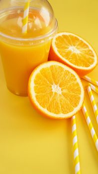 Orange juice in fast food closed cup with tube on yellow background. Sliced orange and yellow paper straws for a drink. Vertical photo