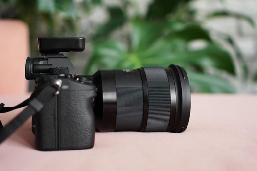 Black camera with a lens on a pink background and a blurred background of green leaves. No visible logos and brands