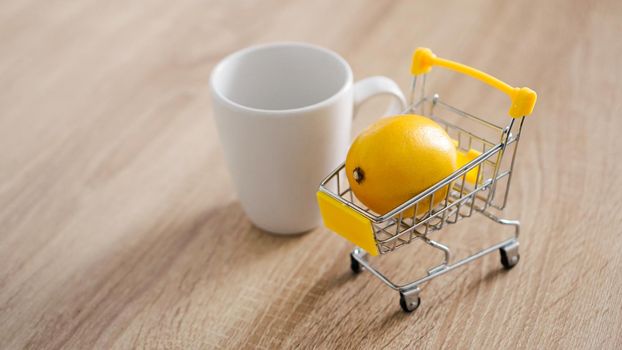 Lemon in a small shopping cart on the kitchen table. Nearby is a white mug of tea. Light and modern kitchen background.