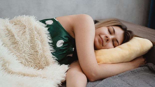 Beautiful young girl in a green nightie sleeps on a yellow pillow on a sofa under a white blanket
