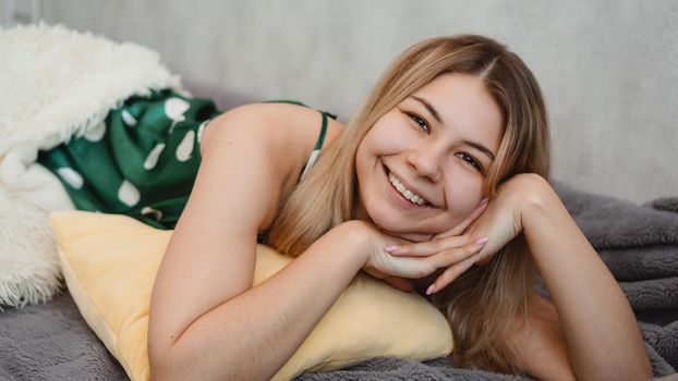 Beautiful smiling blonde in green pajamas on a yellow pillow on a sofa under a white blanket. Good happy morning concept