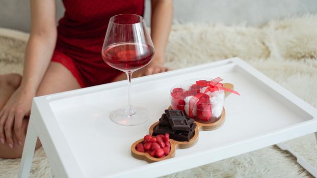 Breakfast for girlfriend. Valentines Day Morning. Wine and sweets on a white tray. Breakfast in bed. Girl in a red peignoir on the background