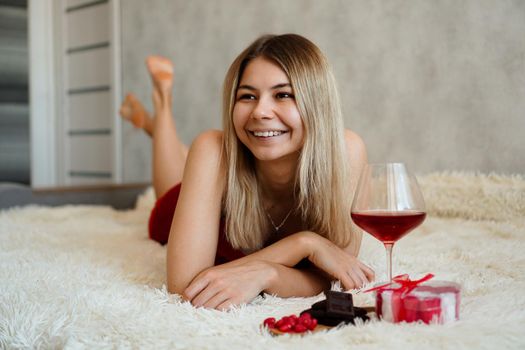 A beautiful smiling blonde lies in bed. Valentines Day Morning. A glass of wine, chocolate, sweets and a gift next to the girl. Happy morning in love