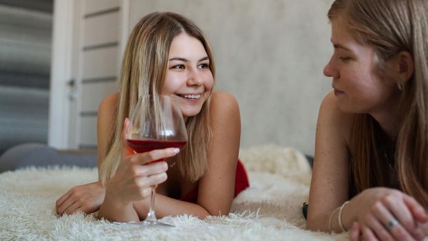 Two friends are lying on the couch and chatting. Beautiful blonde holding a glass of red wine in her hand