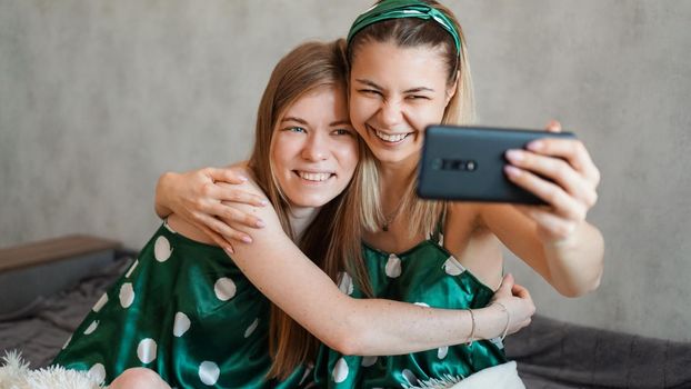Two beautiful happy girlfriends hugging and taking selfies with smartphone at pajama party