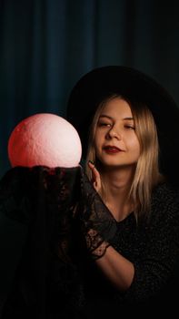 A young woman fortune teller in a hat is holding a magic ball. Beautiful psychic woman is guessing with a red magic ball