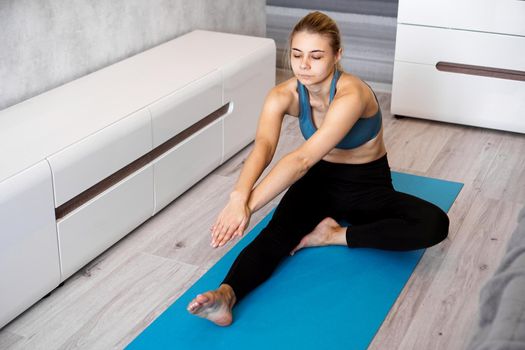 Sport, training and lifestyle concept - woman stretching leg on blue yoga mat at home in the living room