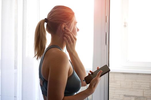 Young pretty sporty girl stands near the window and listens to music on headphones from smartphone. Sunny day